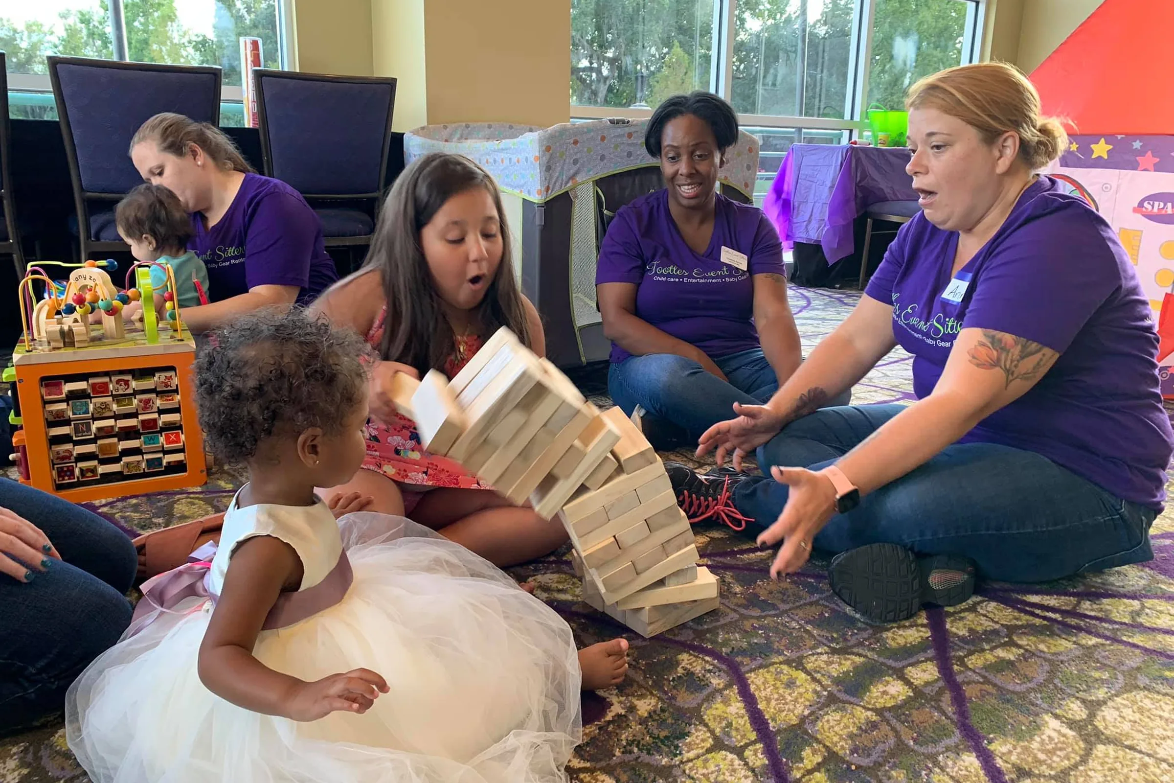 Tootles Event Sitters in branded purple shirts playing a giant block game with children, including a flower girl, in a wedding kids' room at a bright venue